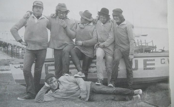 Black and white photograph of Sailing Club in early 1970s. 5 men stand in front of a boat, with one laid on the floor at their feet.